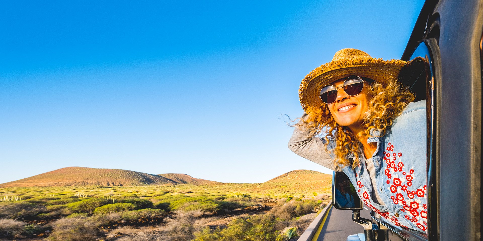 A happy woman in a straw hat and sunglasses hanging out of a car window driving down a paved road in the desert.