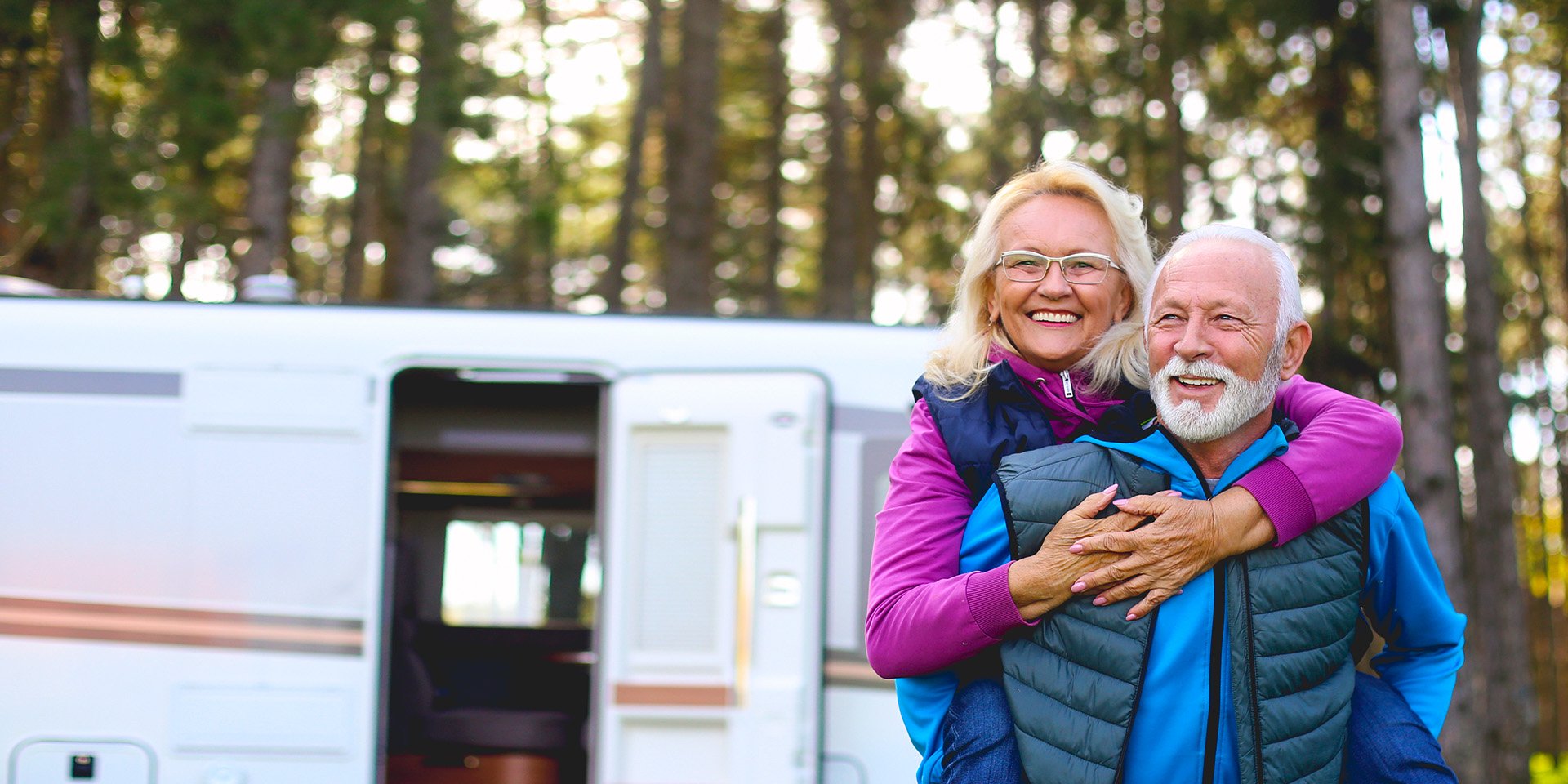 Elderly pair finds joy camping in the woods. The woman rides on the man's back, both smiling, with their RV in the background.