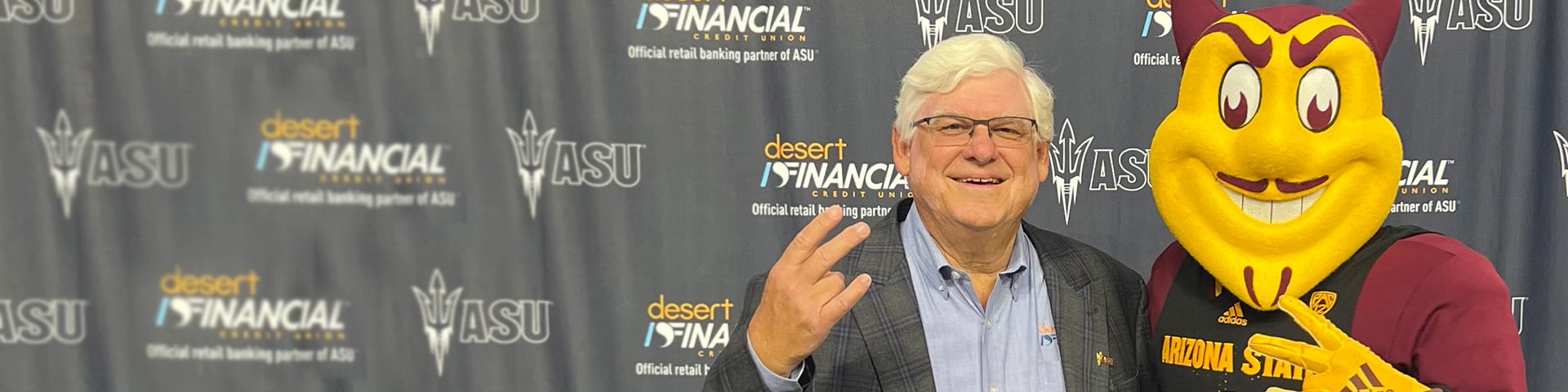 Jeff Meshey (left) proudly displaying the 'Fork 'em Devils' hand sign, standing alongside Sparky the Sun Devil Mascot, against a backdrop celebrating the partnership between Desert Financial and Arizona State University.