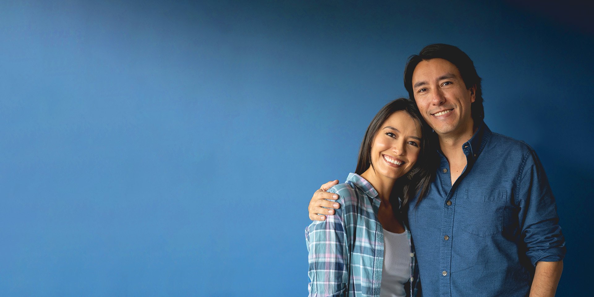 A happy couple hugging each other and smiling for the camera in front of a dark blue background.