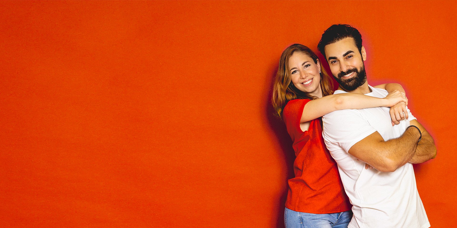 A happy couple standing together in front of a red background as the woman is hugging the man above his shoulders from behind.