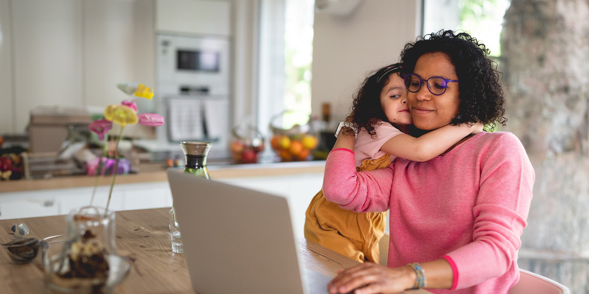 A young daughter embraces her mom warmly at the kitchen table as they find support from Desert Financial, navigating the mortgage assistance process together on their website.