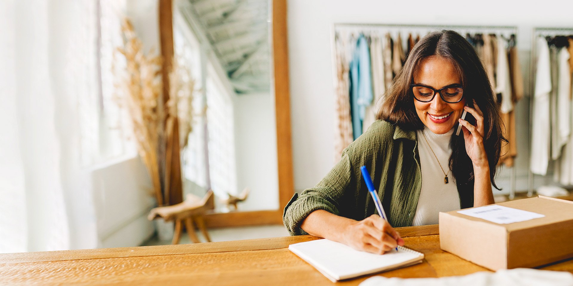 A joyful woman with glasses multitasks on the phone, copying down information into a notebook beside a package on her desk.