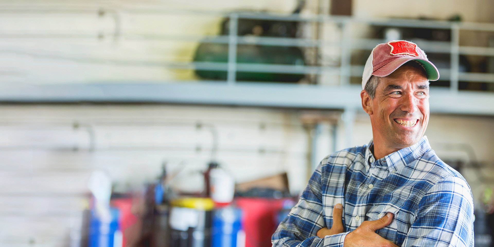 A middle-aged rancher in a ballcap stands confidently in his stable, arms crossed, and smiles off to the side, embodying pride and contentment in his work.