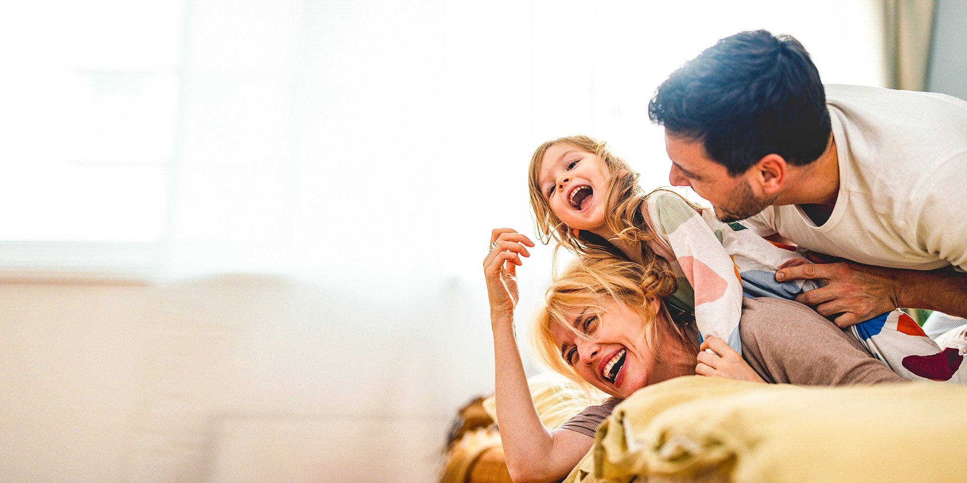 A playful family of three engages in a loving tackle, with the mother at the bottom, the young toddler in the middle, and the father on top, creating a heartwarming and joyous moment.