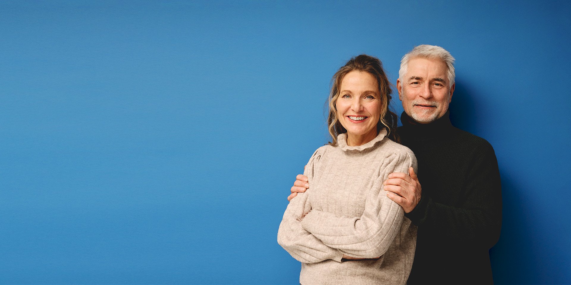 Portrait of an older couple, the wife stands with arms crossed in front of the husband, whose hands rest gently on her shoulders.