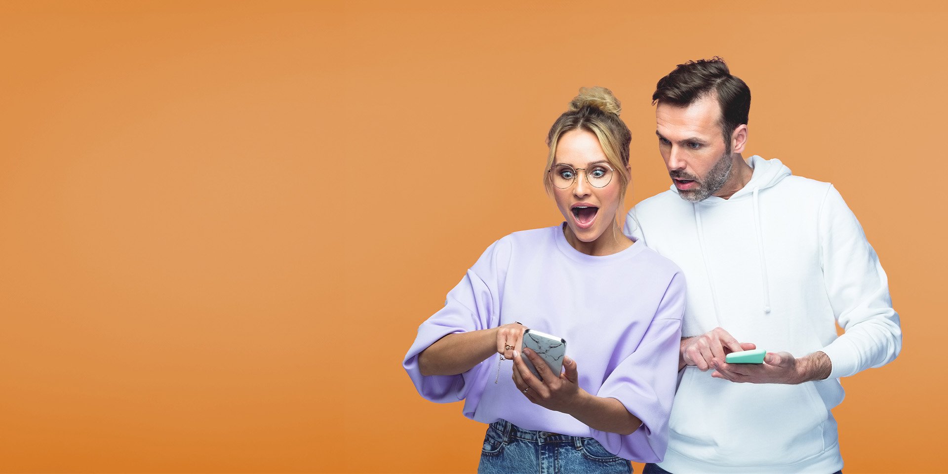 Couple with mobile phones in hand, the woman, with her hair in a bun and wearing glasses, reacts to a surprising screen. The man looks over her phone against an orange background.