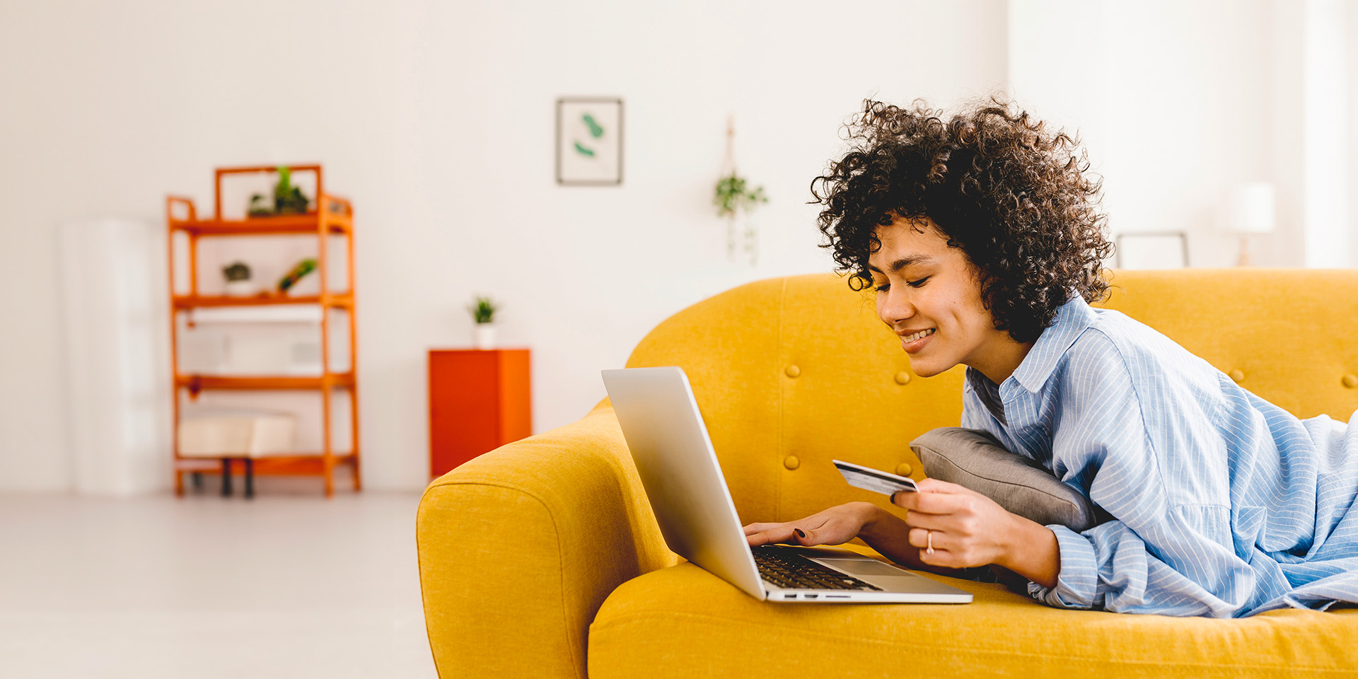 A young woman lies on her stomach on a yellow couch, as she inputs her card information to her laptop. 