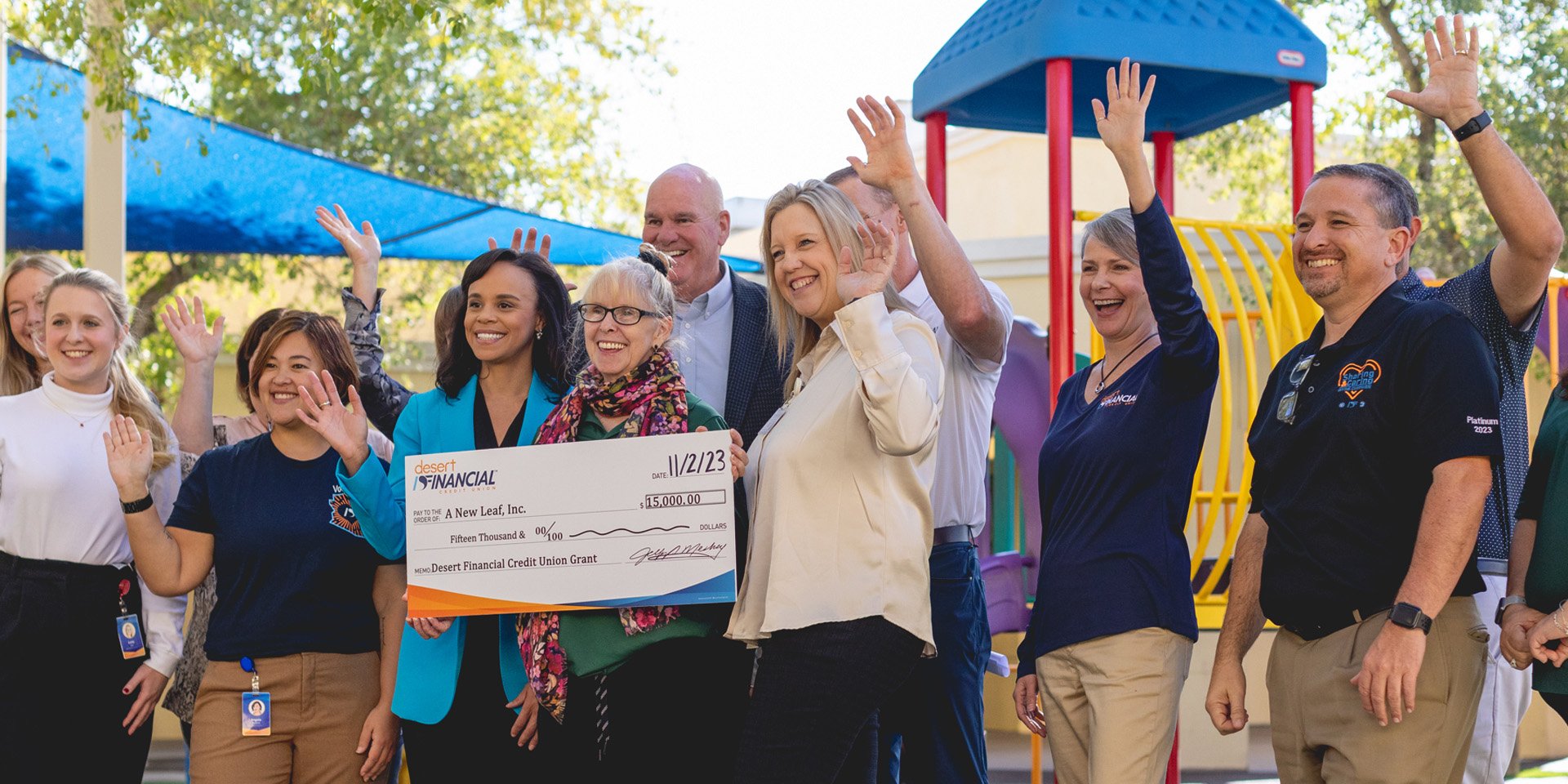 Carlissa Wright and a group of individuals gather around a large check made out to 'A New Leaf, Inc.' for a generous $15,000 grant from Desert Financial. The setting is an outdoor playground, capturing the moment of philanthropy and community support in action.