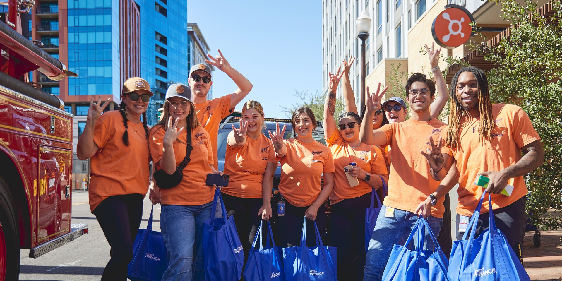 Ten Desert Financial employees and ASU students in orange shirts pose on University Drive in Tempe during homecoming, holding up the 'Fork 'em Devils' hand sign in front of a Tempe Police car.