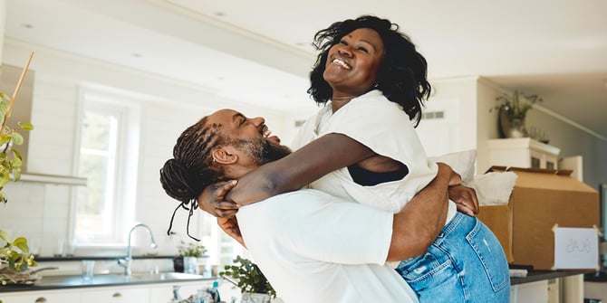 New homeowners celebrate in their kitchen, surrounded by unpacked boxes. Husband joyfully lifts his wife in a warm embrace, marking a moment of happiness in their first home together.