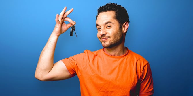 A smirking man dangling car keys up by his head in front of a blue background.