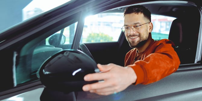 A new graduate sitting in the driver's seat of his car and fixing the side mirror through the window.