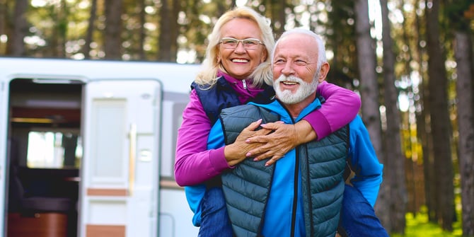 Elderly pair finds joy camping in the woods. The woman rides on the man's back, both smiling, with their RV in the background.
