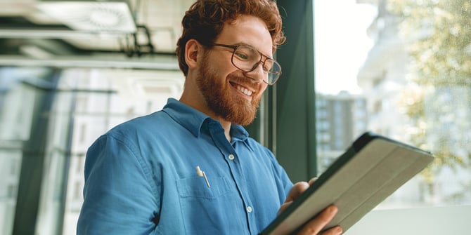 A man with glasses smiles while using the Desert Financial website on a tablet, discovering the benefits of MyMoney@Work for his company and employees, enjoying the positive impact on financial well-being.