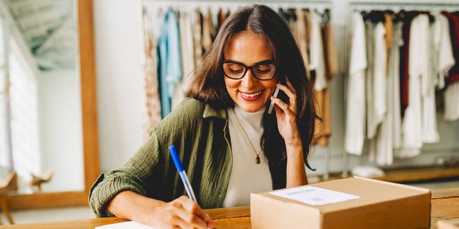 A joyful woman with glasses multitasks on the phone, copying down information into a notebook beside a package on her desk.