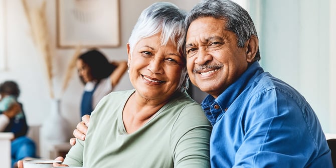 Grandparents embracing each other and posing for the camera as two kids and their mother play in the background.