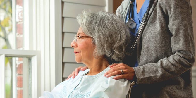 An elderly woman sits on a porch in her wheelchair, with a caring nurse behind, gently pushing her chair.