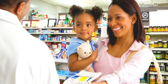 Mother holds daughter, who clutches a teddy bear, while conversing with a pharmacist over the counter at the pharmacy.