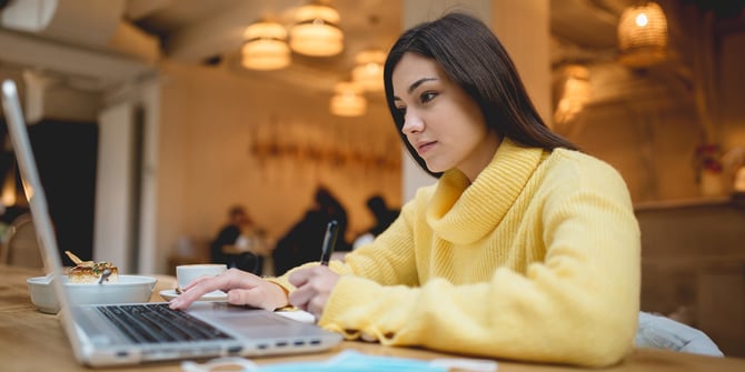 A student in a yellow sweater at a café takes notes from her laptop onto a notebook.