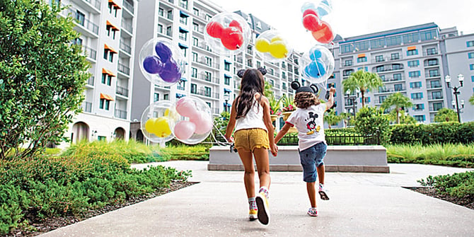 Two joyful children, wearing Mickey Mouse ears, holding hands and skipping away from the camera with a bunch of Mickey Mouse balloons in hand. The scene takes place on the grounds of one of Disney's hotels.