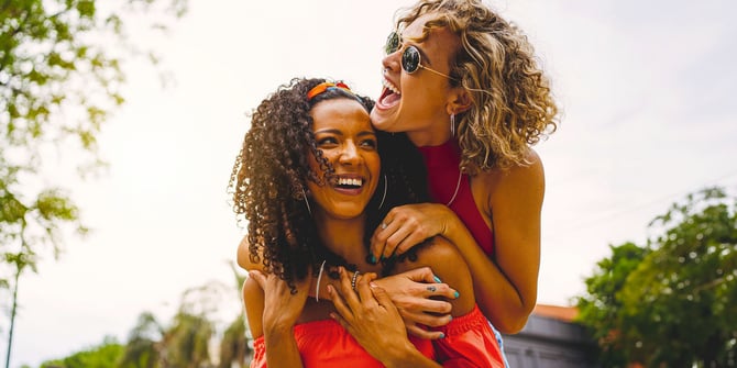 Two joyful women having a great time in an outdoor park. One woman is on the other's back, and both are sharing laughter.