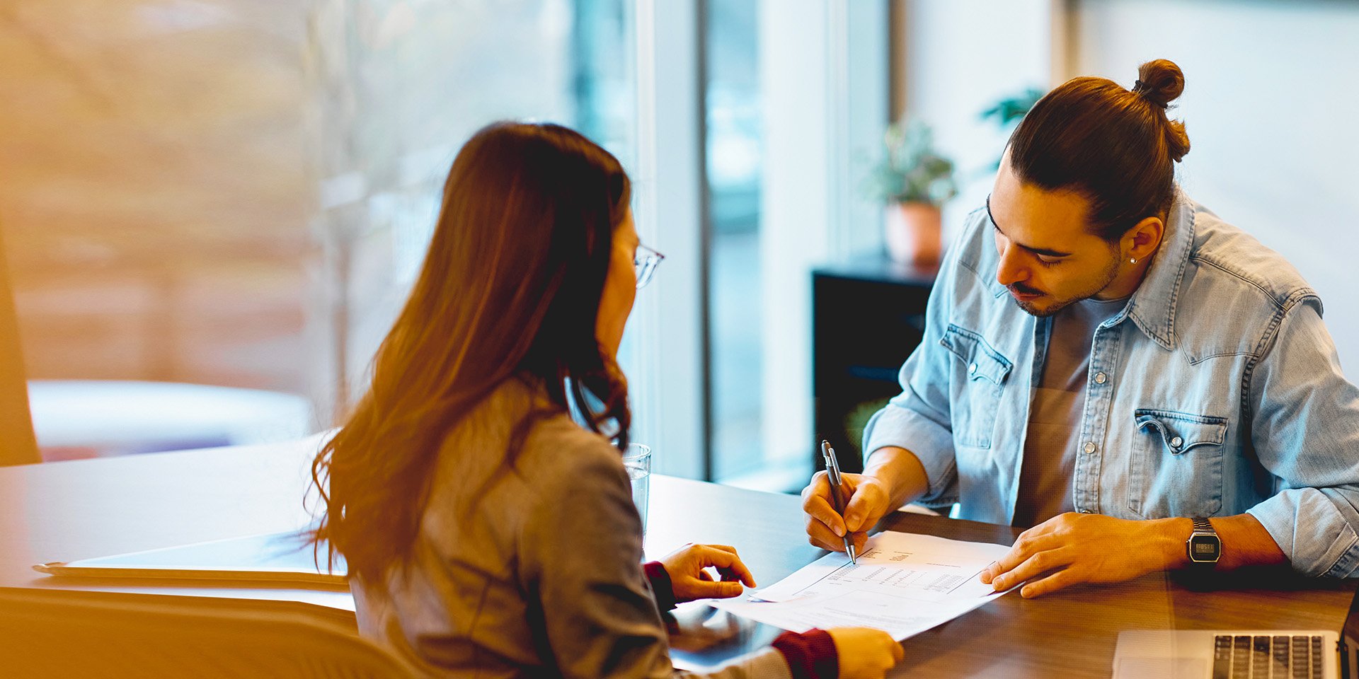 A bank teller helping a young man fill out a written form over the counter. A bank teller helping a young man fill out a written form over the counter.