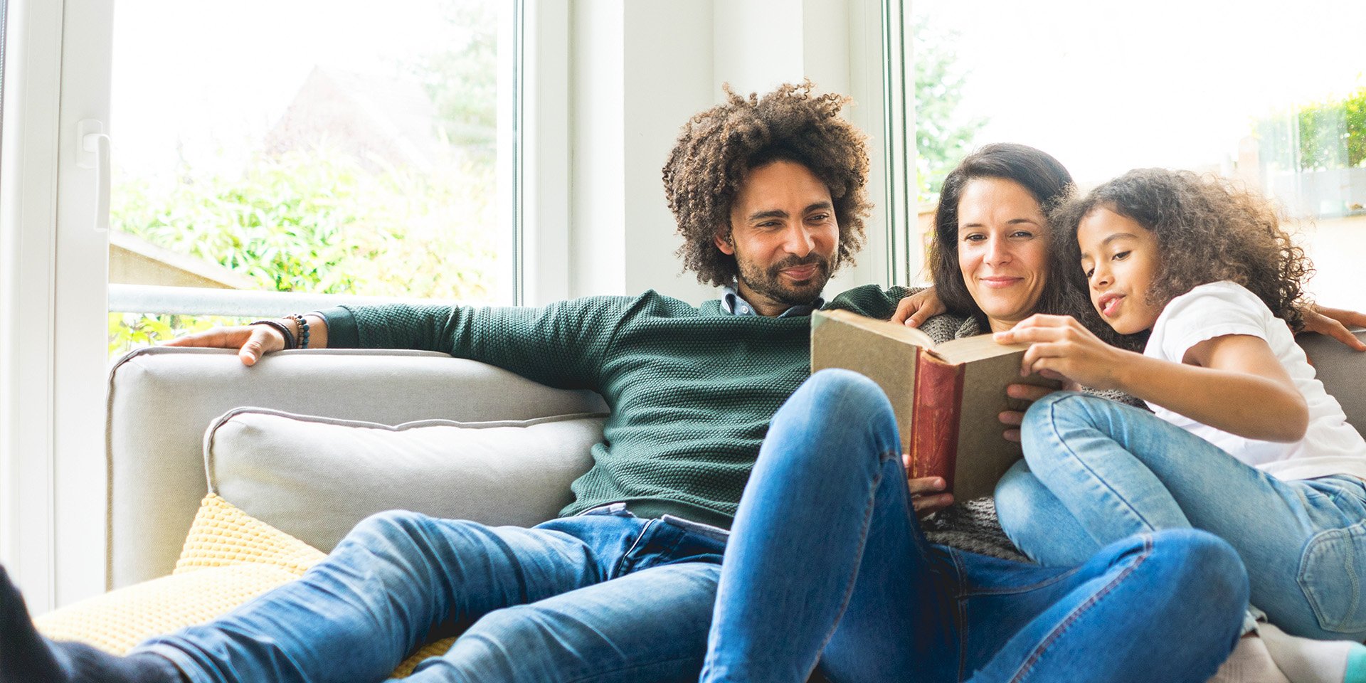 A cozy family moment in their bright living room, with the father, mother, and daughter reading a book together on the couch. The mother holds the book while the young girl eagerly reaches for it.