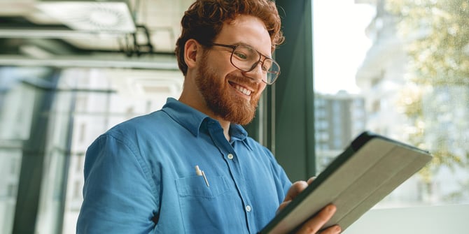 A man with glasses smiles while using the Desert Financial website on a tablet, discovering the benefits of MyMoney@Work for his company and employees, enjoying the positive impact on financial well-being. A man with glasses smiles while using the Desert Financial website on a tablet, discovering the benefits of MyMoney@Work for his company and employees, enjoying the positive impact on financial well-being.