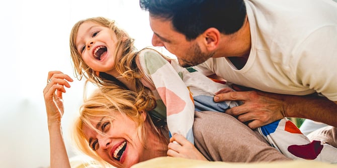 A playful family of three engages in a loving tackle, with the mother at the bottom, the young toddler in the middle, and the father on top, creating a heartwarming and joyous moment.