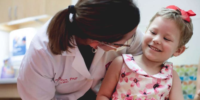 A compassionate Phoenix Children's Hospital Pediatric Nurse Practitioner, wearing a mask and glasses, comforts a young girl in her exam room. The nurse practitioner gives the girl a warm hug, creating a reassuring and joyful moment that brings a smile to the child's face.