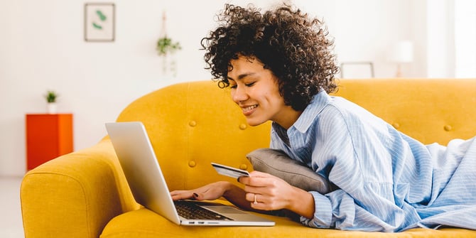 A young woman lies on her stomach on a yellow couch, as she inputs her card information to her laptop. 