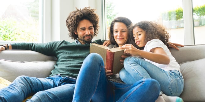 A cozy family moment in their bright living room, with the father, mother, and daughter reading a book together on the couch. The mother holds the book while the young girl eagerly reaches for it.